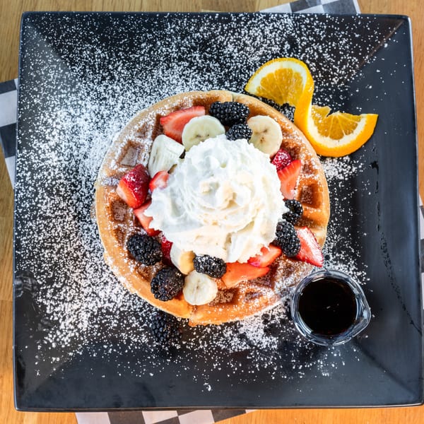 Golden waffle topped with whipped cream, strawberries, bananas, blackberries, and powdered sugar, with syrup and orange slices.