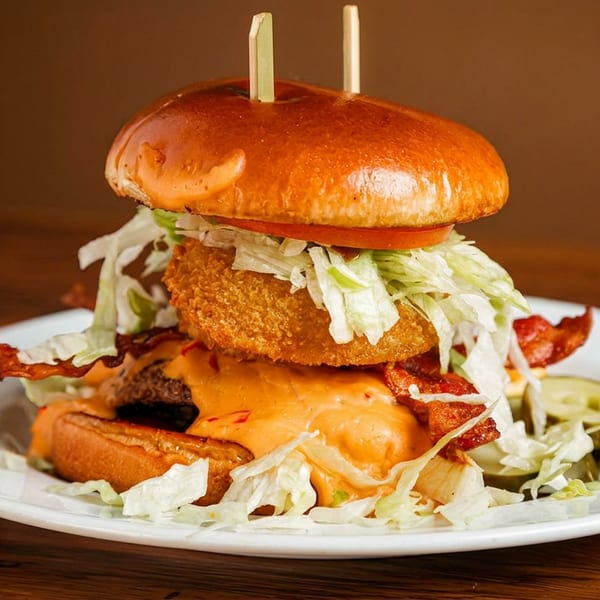 A loaded burger with melted cheese, crispy bacon, shredded lettuce, tomato, and a fried onion ring.