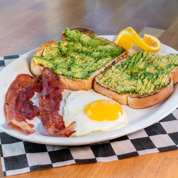 Breakfast plate with avocado toast, crispy bacon, a fried egg, and orange slices on a checkered napkin.