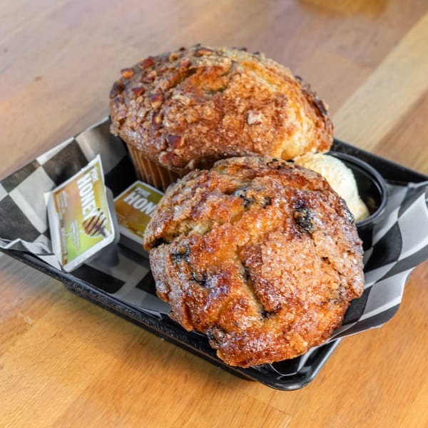 Two muffins, one blueberry and one nut-topped, with butter and honey packets on a checkered tray.
