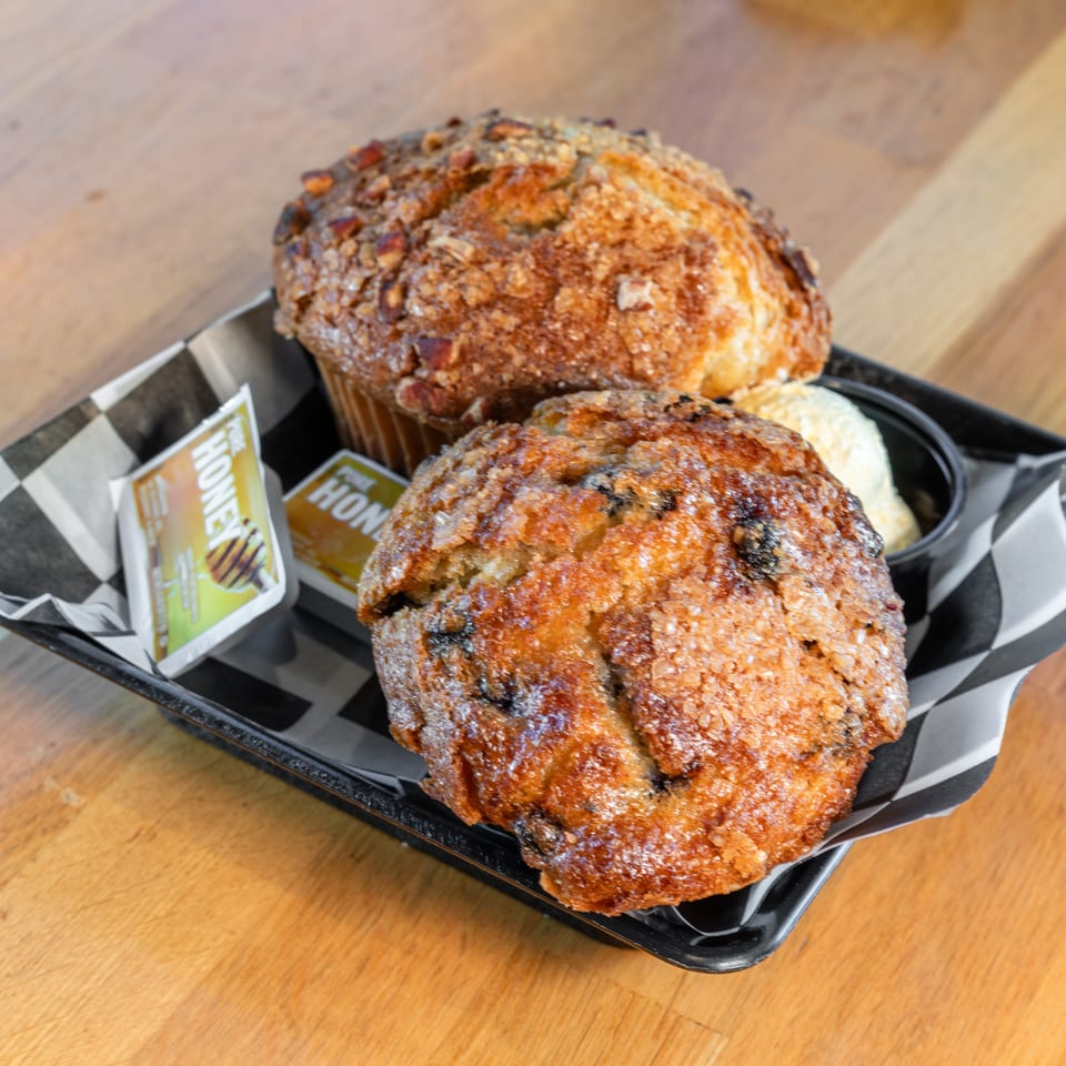 Two muffins, one blueberry and one nut-topped, with butter and honey packets on a checkered tray.