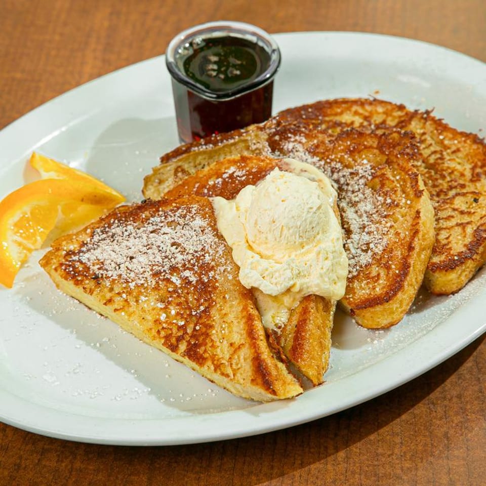 Golden French toast topped with ice cream, powdered sugar, syrup, and an orange slice.
