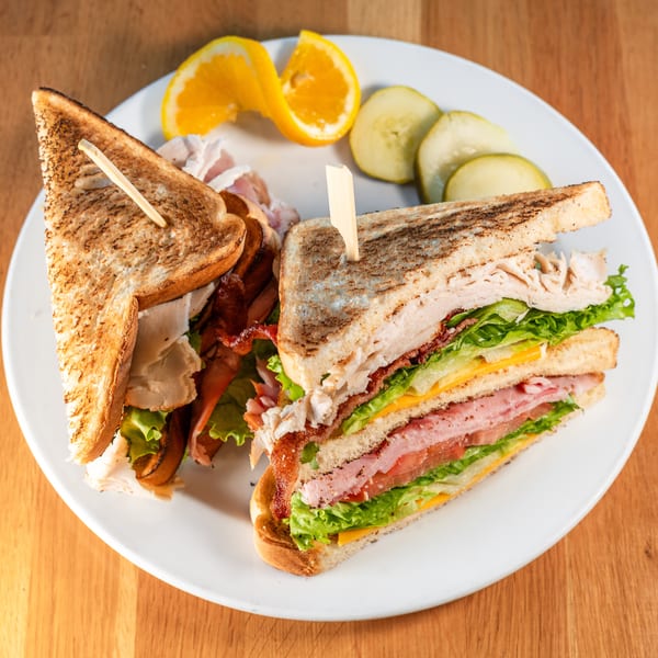 A club sandwich with turkey, ham, bacon, lettuce, tomato, and cheese, served with pickles and orange slices.