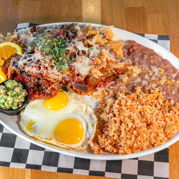 A large plate of chilaquiles with two fried eggs, rice, refried beans, guacamole, and orange slices.
