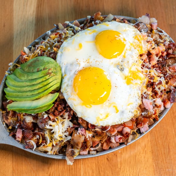 Breakfast skillet with two fried eggs, avocado slices, bacon, ham, and cheese on a wooden table.