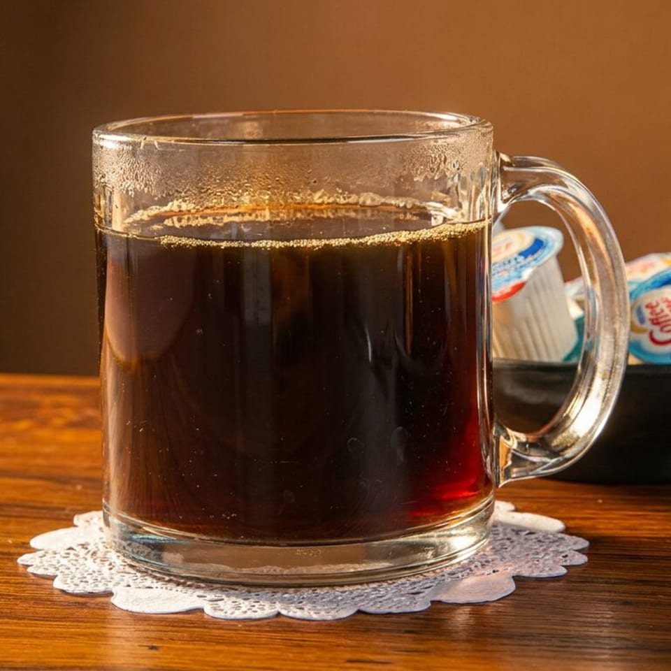 A glass mug of hot coffee on a lace doily, with creamer packets in a bowl beside it.