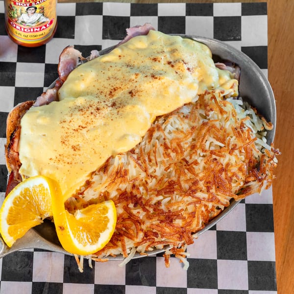 Eggs Benedict with hash browns, two orange slices, and hot sauce bottles on a checkered paper.