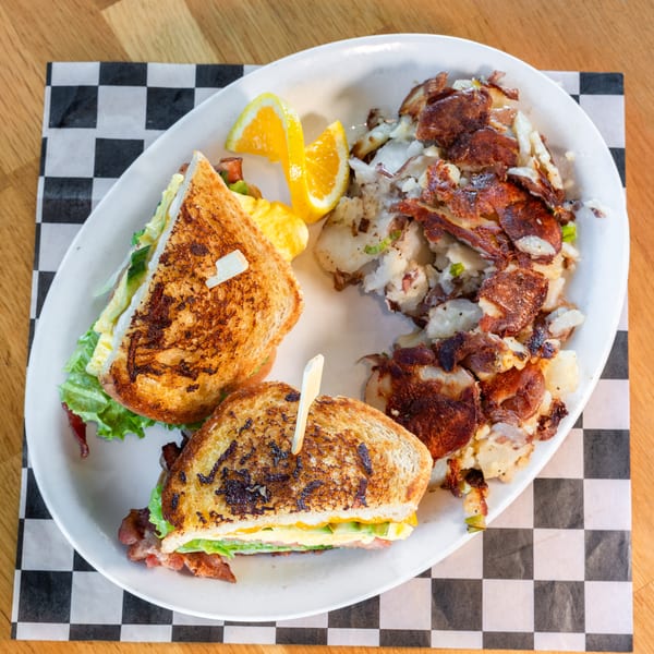 Breakfast sandwich with egg, bacon, and lettuce, served with home fries and orange slices.